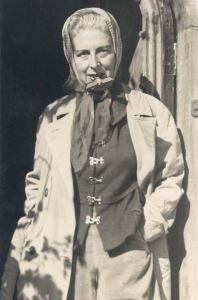 Claude Cahun as an older woman, standing at the door of her home during the liberation of Jersey, defiantly holding a Nazi badge between her teeth