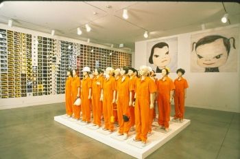 MOCA Gallery's photograph of a room of the 2001 Superflat exhibition shows groovision's Chappies, standing in their orange uniforms in the center of the room, and two works by Yoshimoto Nara behind them.