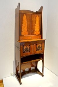 Joseph Maria Olbrich's <i>Cupboard</i> (1900), made of stained maple, has colored inlays that replicate Art Nouveau motifs, though its curved lines in the legs and at the top also contribute to the piece's aesthetic elegance.